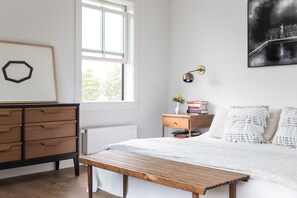 Ein Schlafzimmer mit einer hölzernen Bank, einem Nachttisch mit Büchern und einer Vase, einem Heizkörper und einem Fenster mit Jalousien.