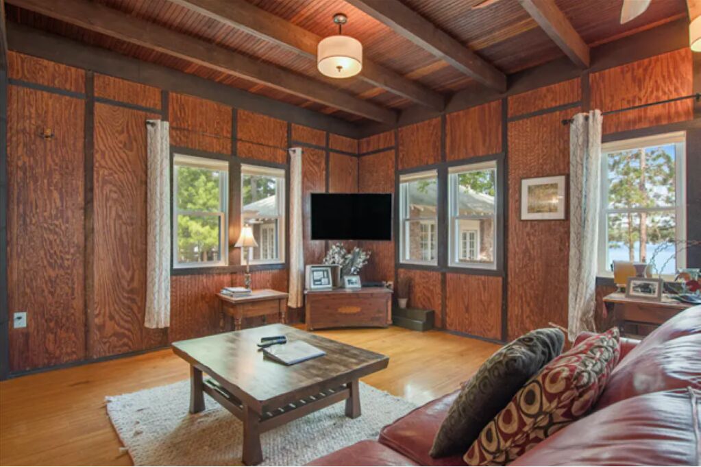 A spacious living room with wooden paneling, large windows, a flat-screen TV, a wooden coffee table, and a leather sofa.