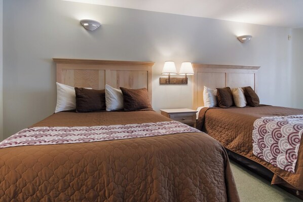 A hotel room with two beds, each with a brown quilted bedspread and a patterned throw. There are two wall-mounted lamps between the beds.