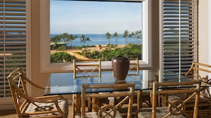 Un comedor con una mesa de vidrio y sillas de mimbre, con vista a una playa con palmeras y el océano.