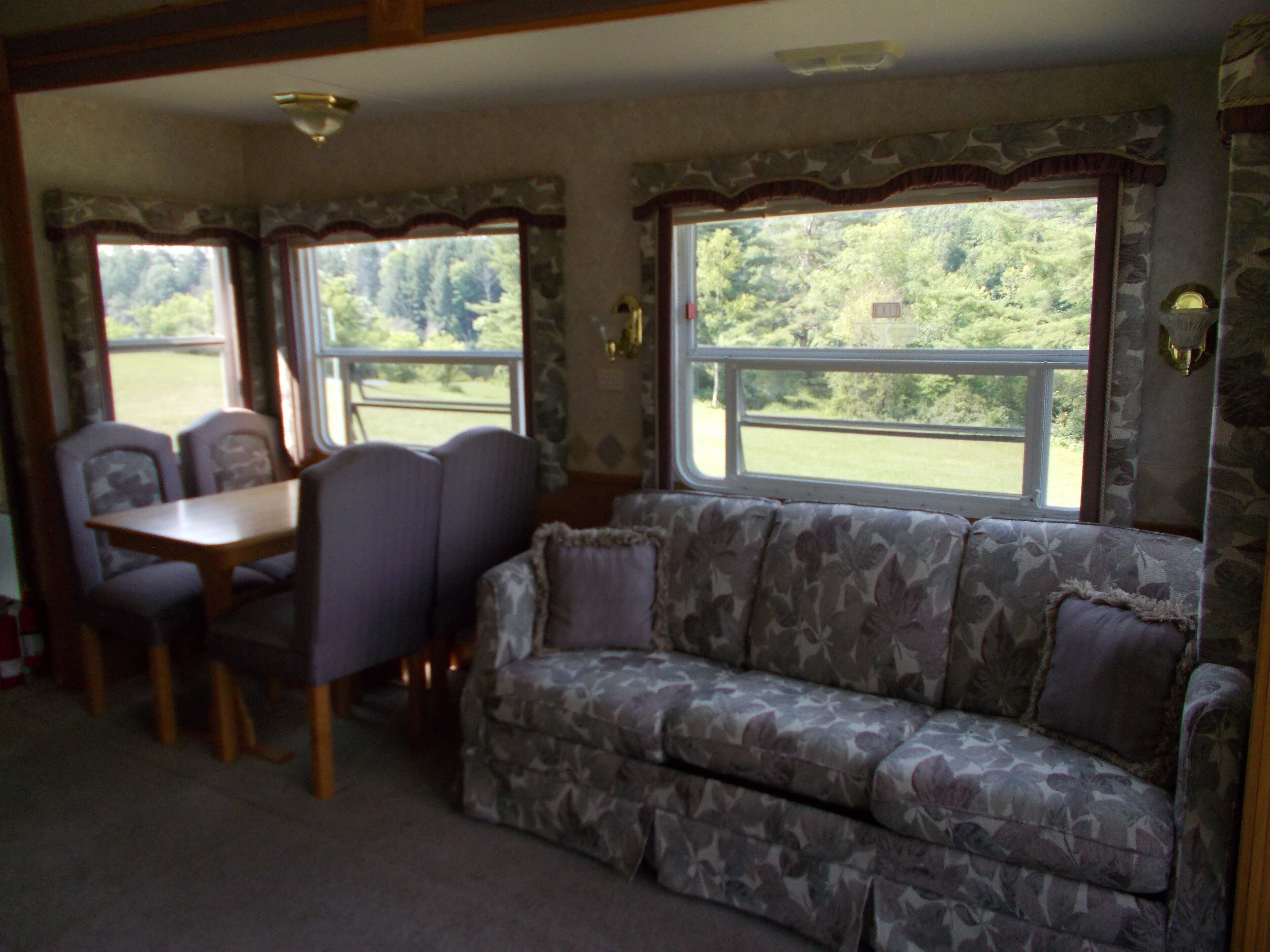 Ein gemütliches Wohnmobil-Interieur mit Esstisch und Stühlen, einer blumengemusterten Couch und großen Fenstern mit Blick auf viel Grün.