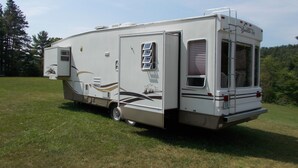 A stationary recreational vehicle (RV) with a slide-out awning, a small enclosed kitchen area, and a rear door with a window. - Private RV Camping on the Connecticut River with local mountain views!! (Monroe)