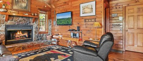 A cozy cabin interior with a fireplace, leather seating, a mounted television, and wooden walls.