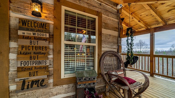 A rustic wooden cabin with a welcoming sign, a wicker chair, and a view of the woods. - AUTHENTIC ANTIQUE LOG CABIN LOCATED IN TIMBERIDGE COMMUNITY (Gatlinburg)