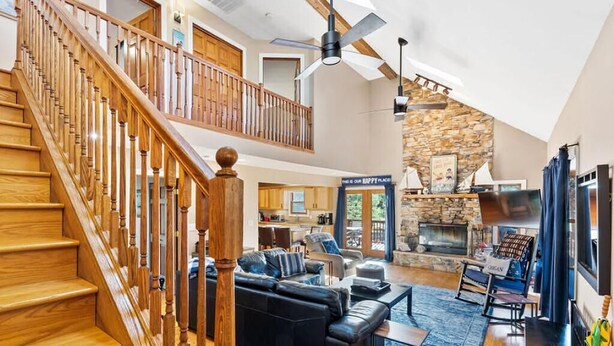A spacious living area with a wooden staircase, a stone fireplace, and a modern kitchen in the background.