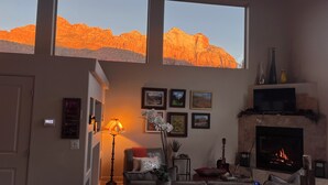 A living room with a fireplace, a sofa, a coffee table, and a TV. There are framed pictures on the wall and a window with a view of mountains. -  Beautiful Home with private Infinity Edge Pool 2 miles to ZNP main entrance (Springdale)