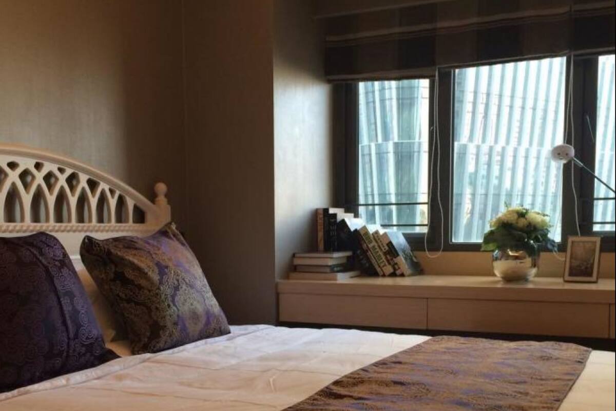 Een netjes opgemaakt bed met een beddekleed met patroon, een hoofdbord en kussens. Er is een raam met jaloezieën, een nachtkastje met boeken en een bloevas, en een lamp.