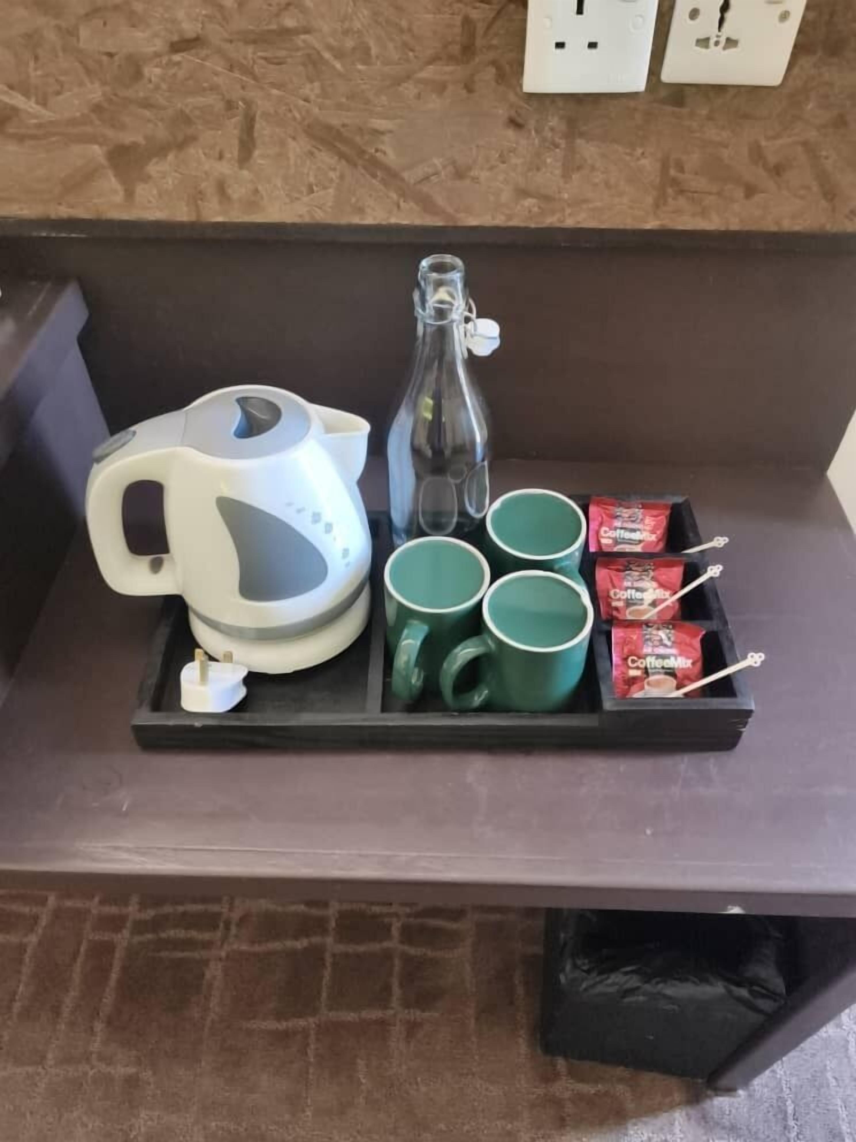 Ein Tablett mit einem weißen Wasserkocher, einer Glasflasche, grünen Tassen und roten Kaffeebeuteln auf einer dunklen Arbeitsplatte.