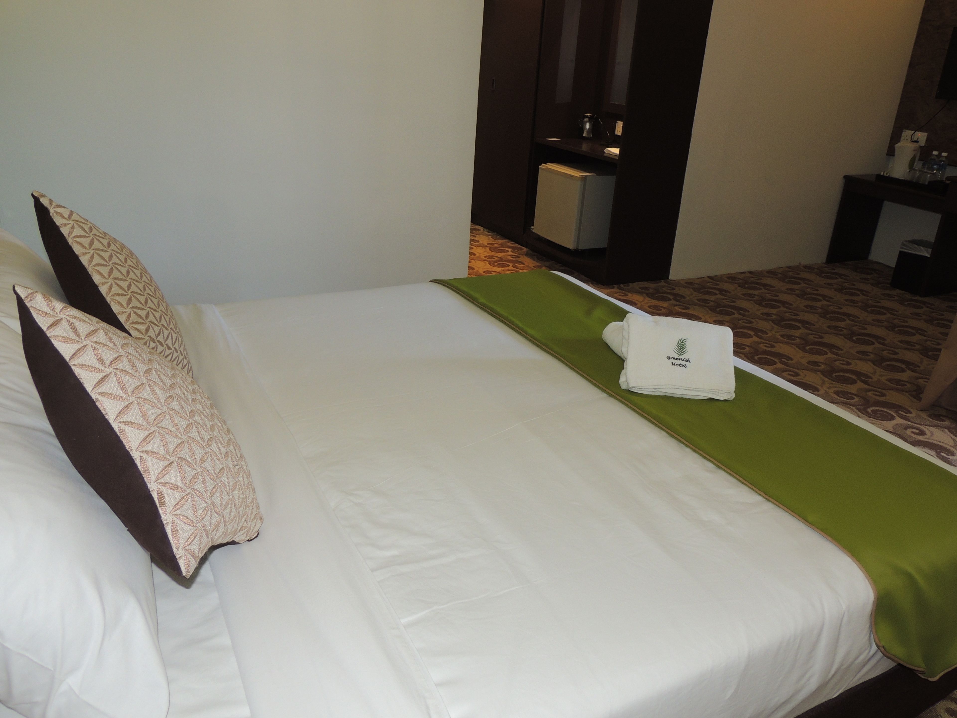 A hotel room with a bed, pillows, a green bedspread, and a towel with the hotel logo.