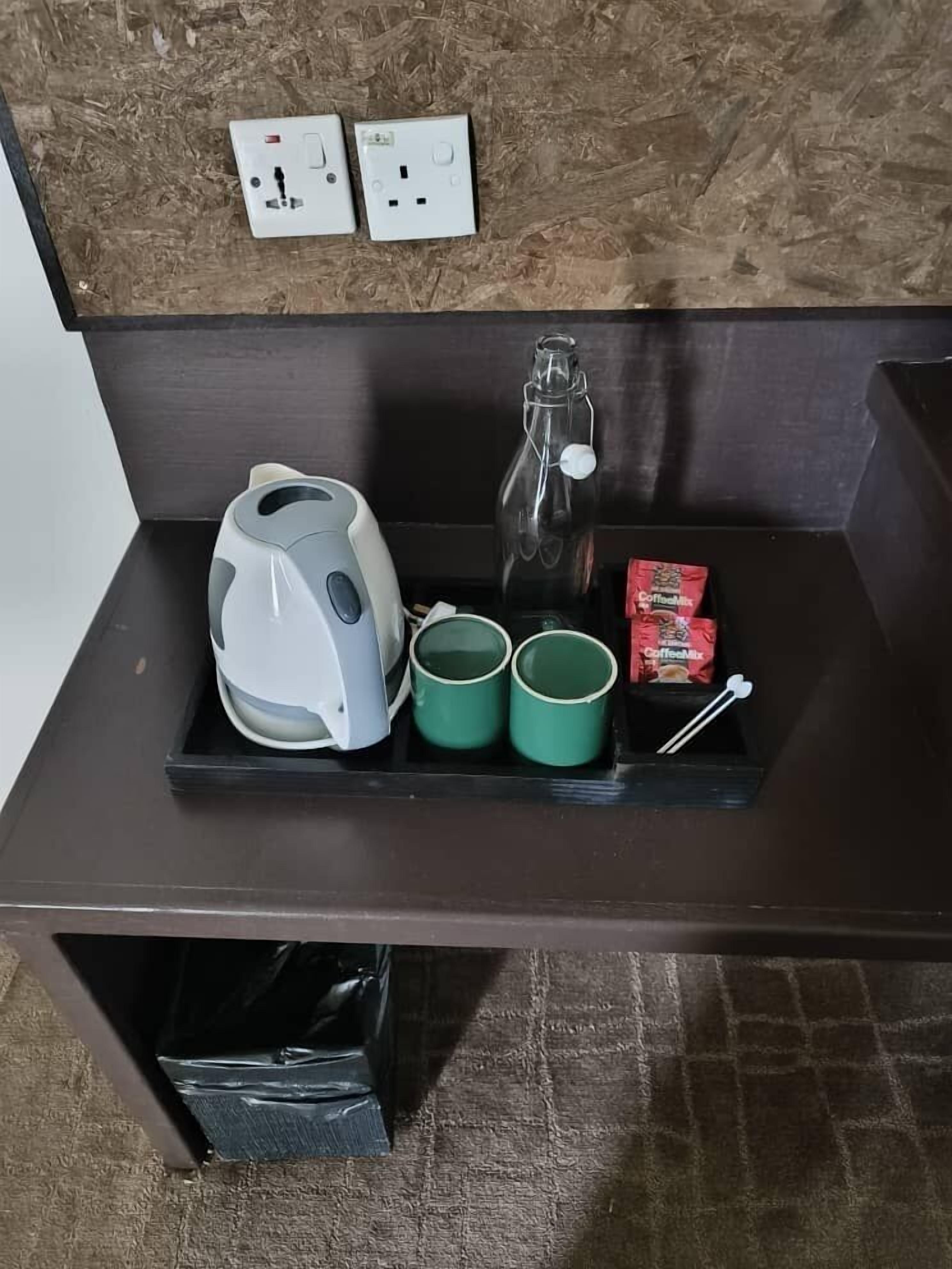 A kitchenette with a kettle, two mugs, a bottle, and a packet of coffee mix on a dark countertop.