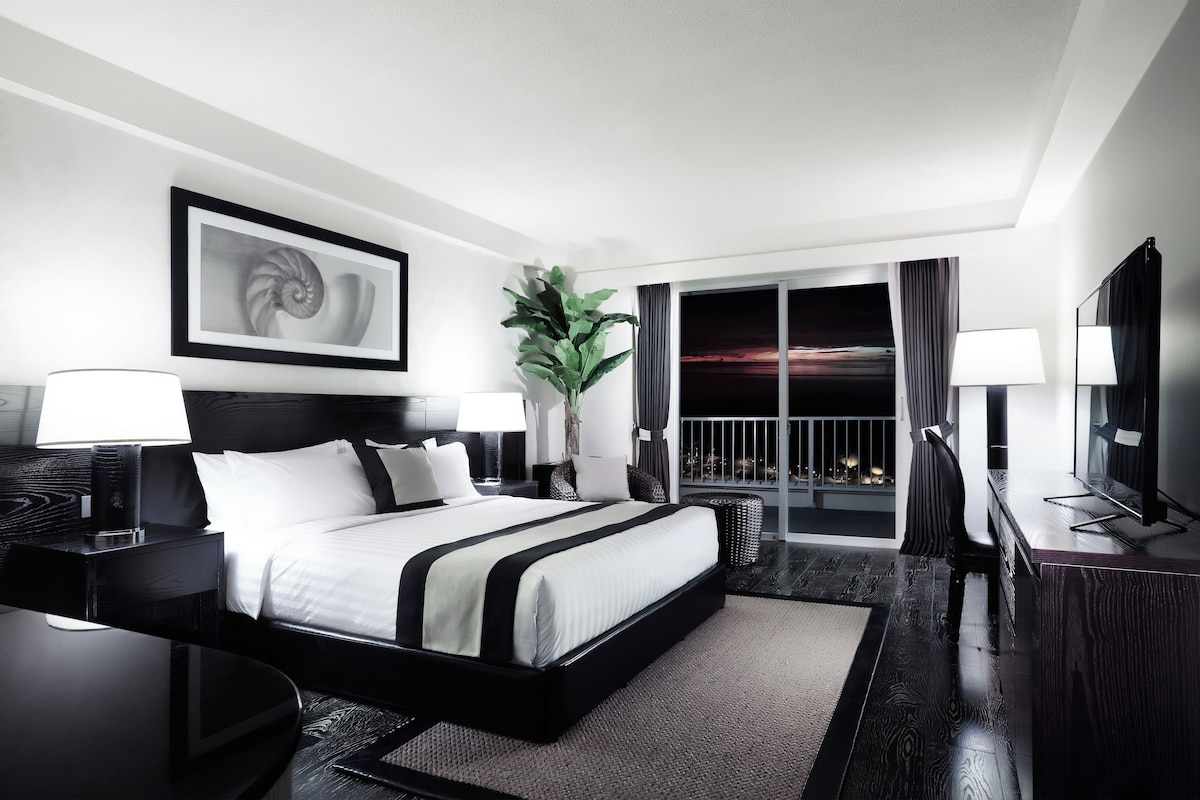 A modern hotel room with a large bed, two bedside tables with lamps, a large window with a view of the city at night, and a framed artwork above the bed.