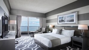 A hotel room with a large bed, a TV, a desk, and a view of the ocean.