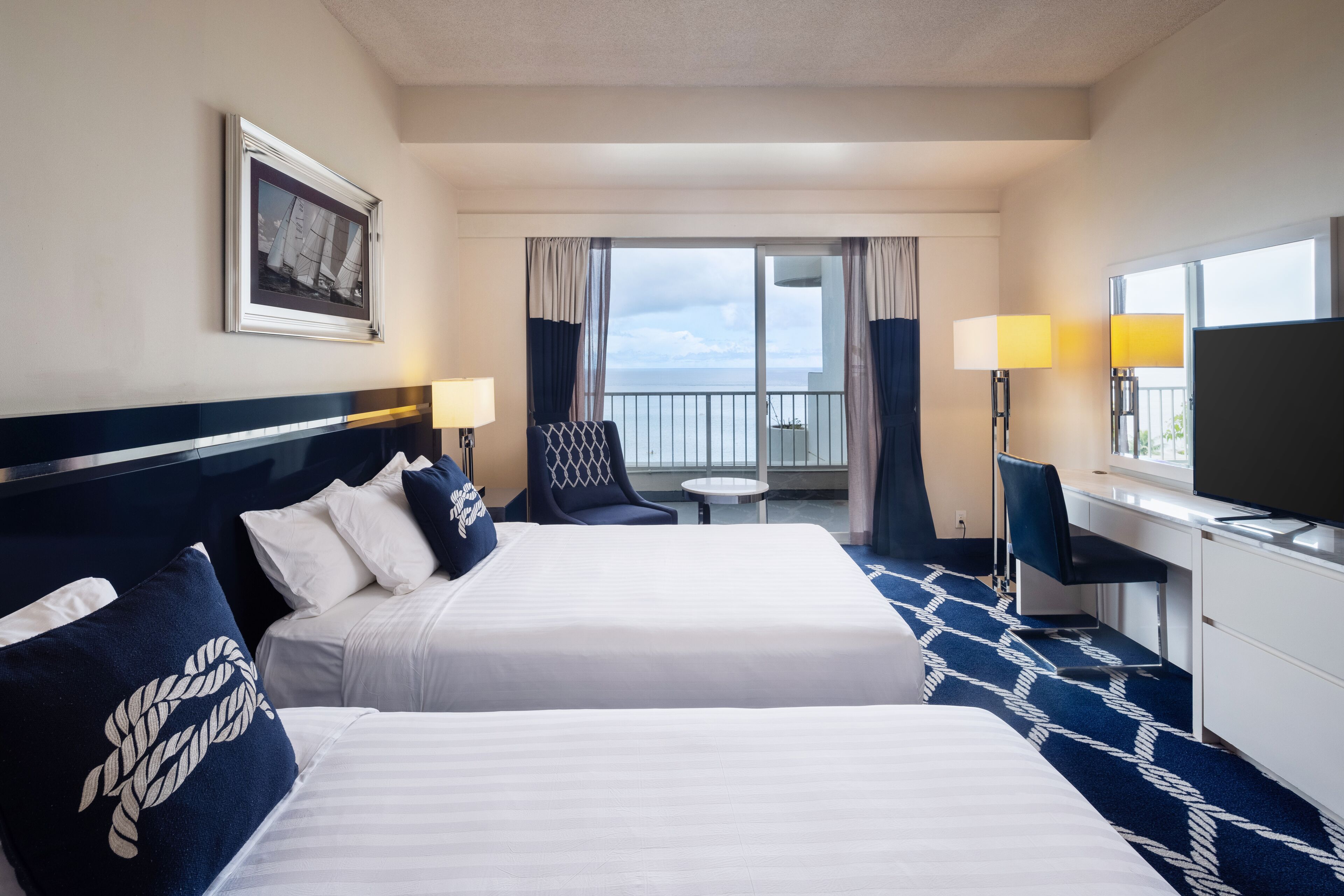 A hotel room with two beds, a desk, a chair, a TV, and a balcony with a view.