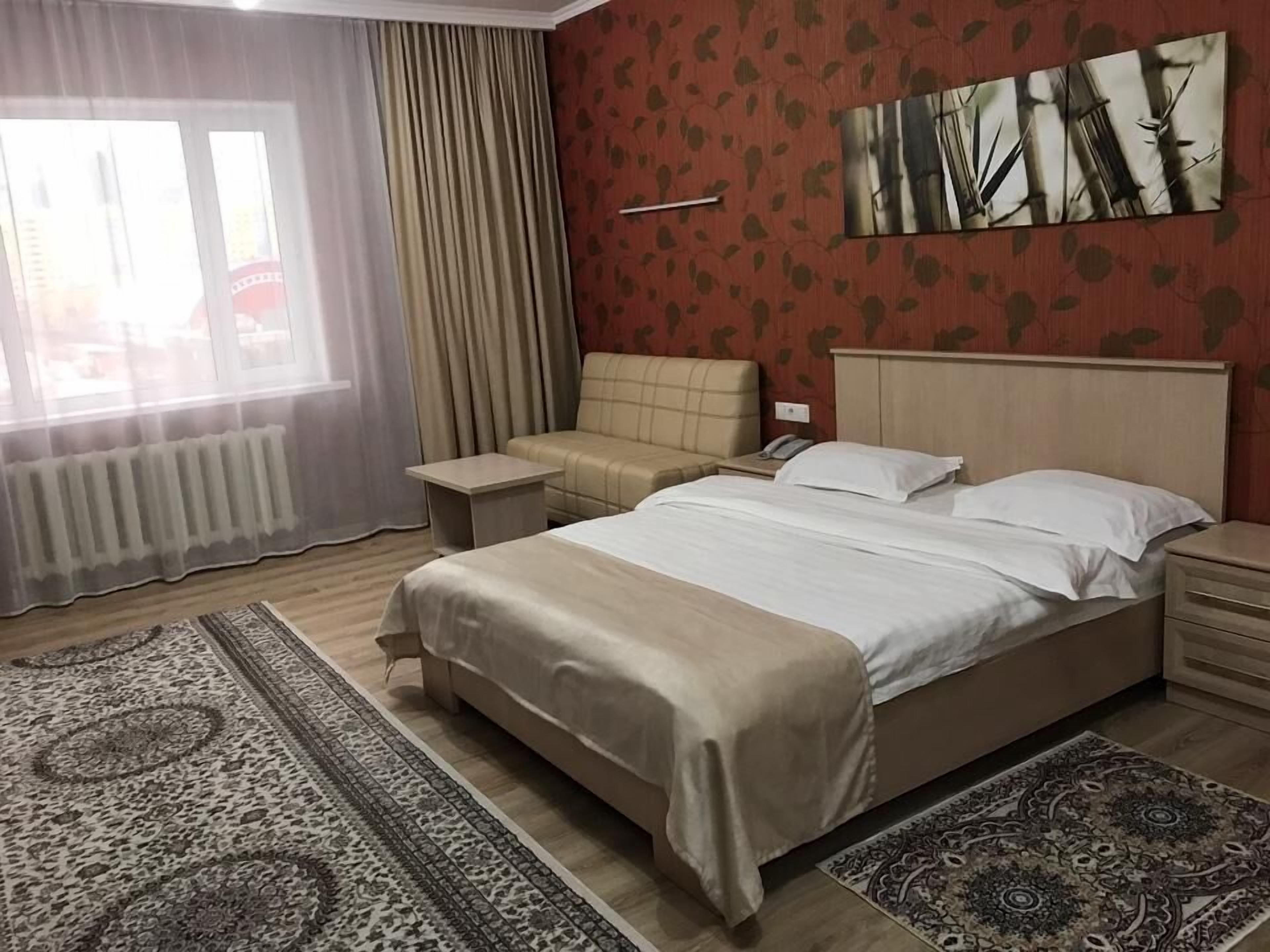 A hotel room with a large bed, a sofa, a nightstand, a bedside table, a radiator, a window with curtains, and a patterned rug.