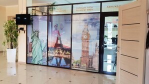 Een lobby met een grote glazen wand waarop bekende bezienswaardigheden staan afgebeeld, een flatscreen televisie en een kamerplant.