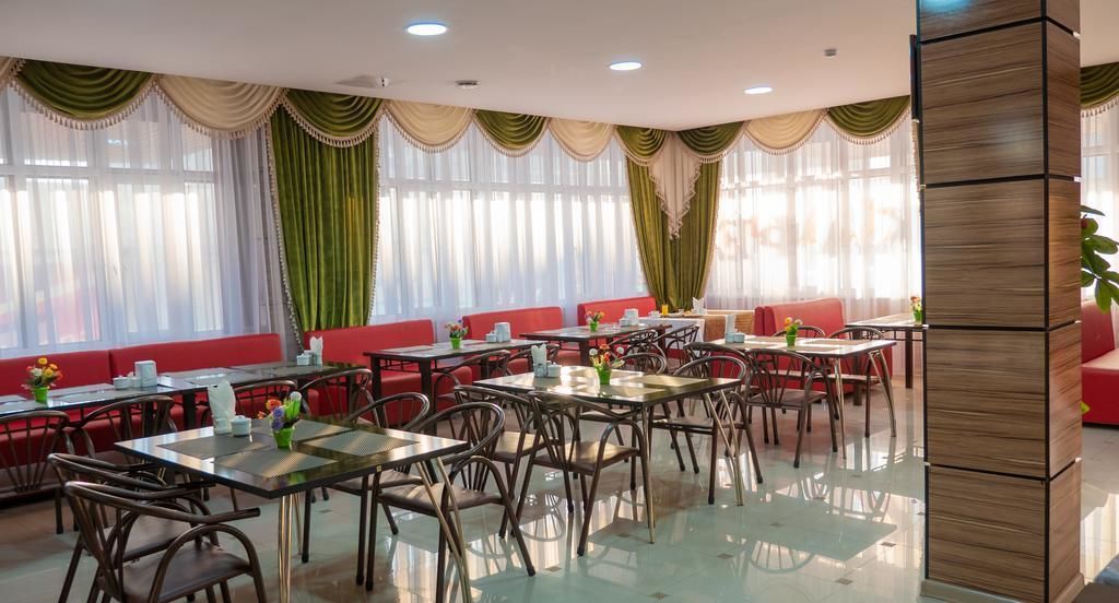 A restaurant with multiple tables and chairs, red cushioned seating, and large windows with sheer curtains.