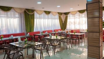 A restaurant with multiple tables and chairs, red cushioned seating, and large windows with sheer curtains.