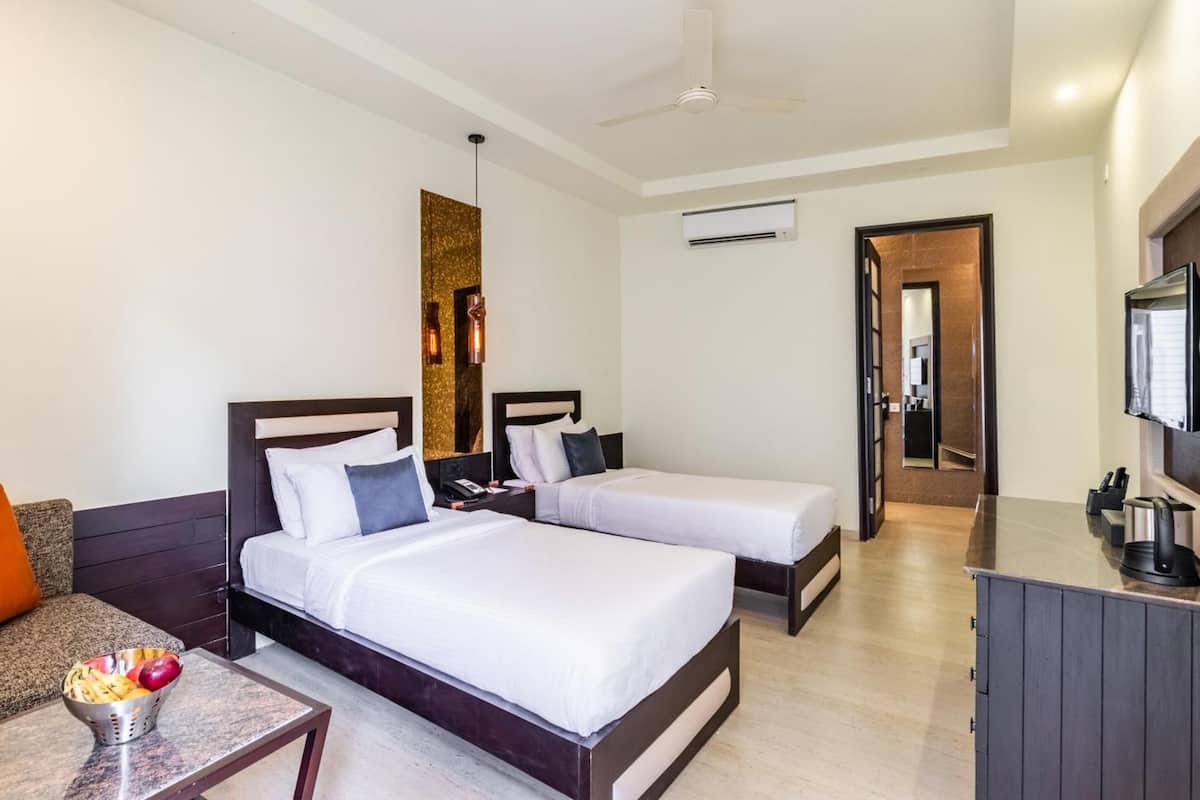 A hotel room with two beds, a sofa, a small table with fruit, a TV, and a minibar.