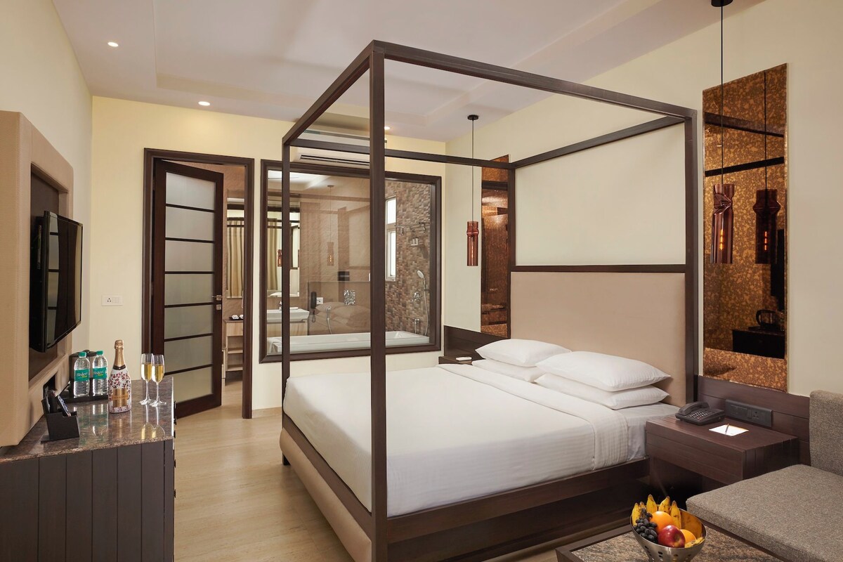 A modern hotel room with a four-poster bed, a television, a minibar, and a fruit bowl on the side table.