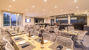 A spacious restaurant with multiple dining tables and wicker chairs, large windows, and a modern kitchen area. - Acacia Resort & Spa Morjim Goa (Morjim)