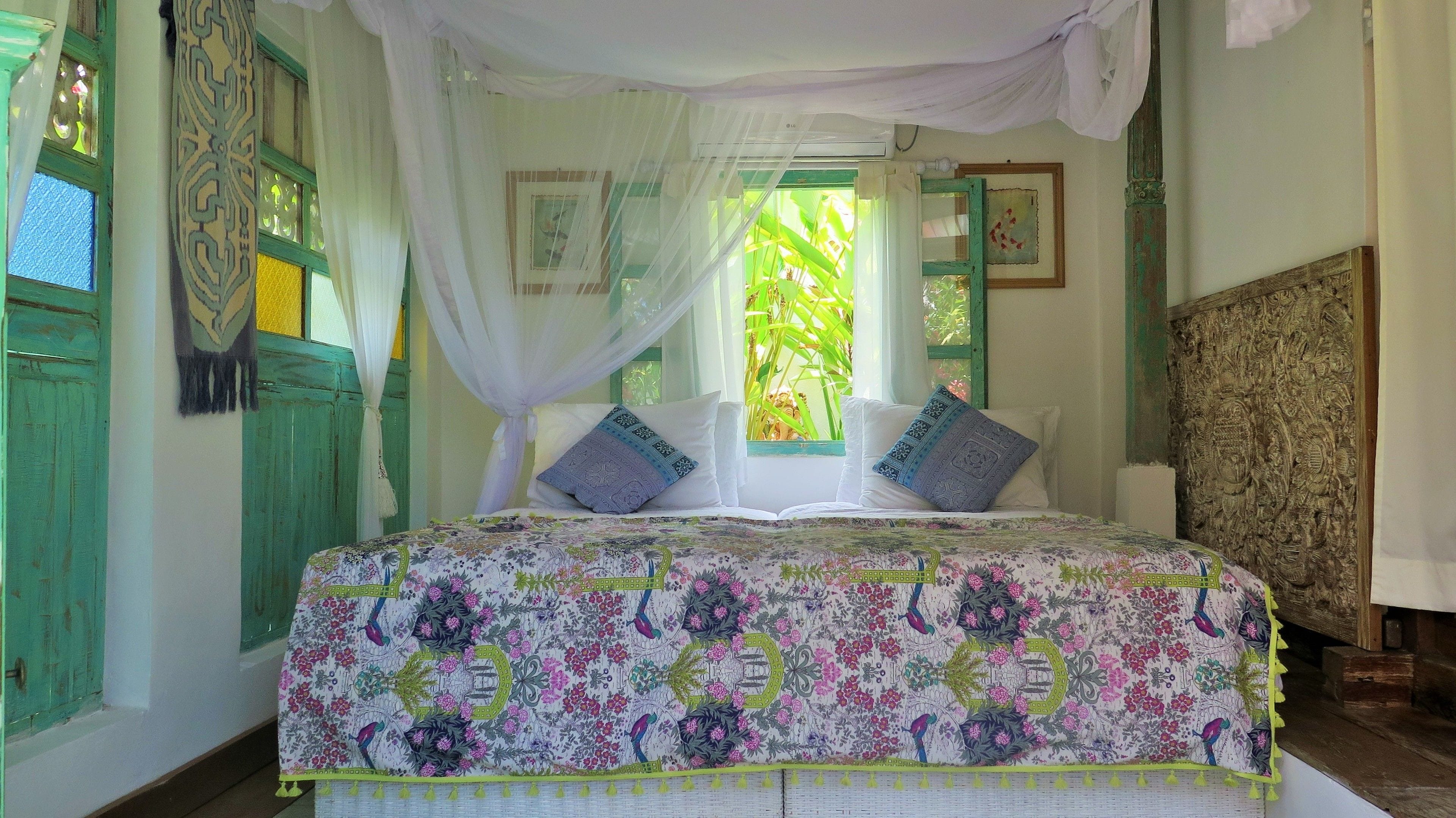 A room with a bed, green shutters, a window with a view of greenery, and a decorative headboard.
