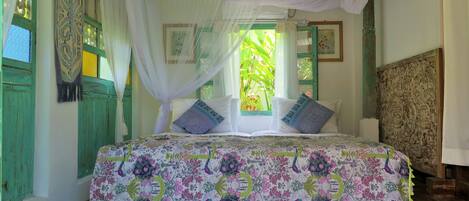 A room with a bed, green shutters, a window with a view of greenery, and a decorative headboard.