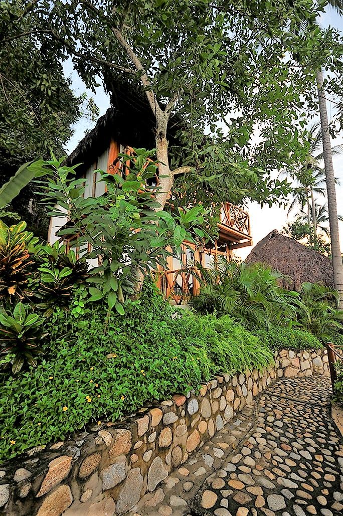 Un sendero de piedra conduce a un edificio tradicional con techo de paja, rodeado de abundante vegetación y una estructura de madera.