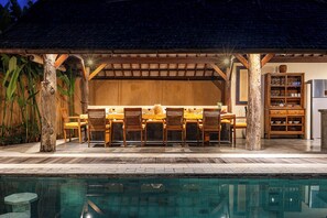 Un espace extérieur aménagé pour les repas, avec des chaises et des tables en bois, une piscine et une structure en bois surmontée d’un toit en tuiles.