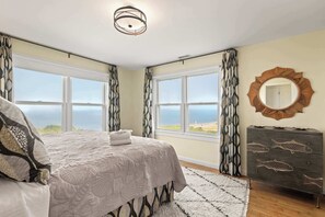 A bedroom with a bed, a dresser, a mirror, and two windows with a view of the sea. - Gorgeous Architectural Waterfront Property! (Eastham)