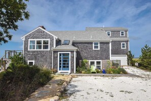 A two-story house with a grey shingle exterior, a blue front door, and a small porch area with a green chair and a blue object on the ground. - Gorgeous Architectural Waterfront Property! (Eastham)