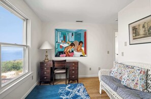 Een kamer met uitzicht op zee, een bureau met lades, een stoel, een bank met gestreepte kussens en een schilderij aan de muur.