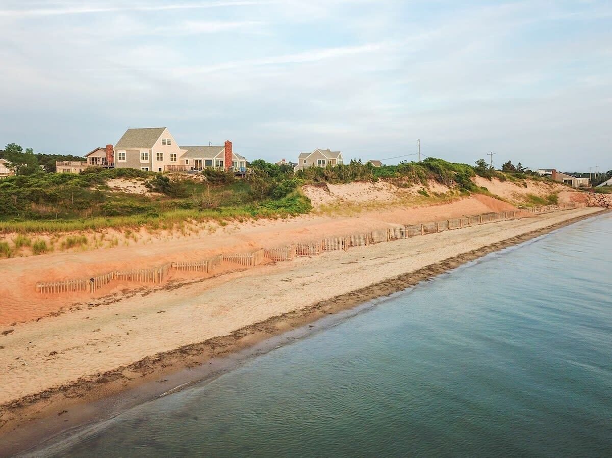 Une propriété située sur la côte, dotée d’une plage de sable, d’une clôture en bois et d’une grande maison avec un toit en pente.