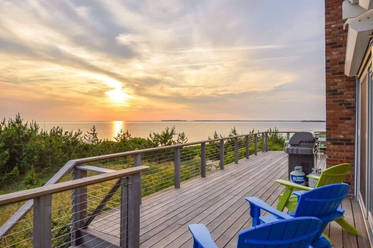 A deck with a view of the sunset over water, a grill, and outdoor seating.