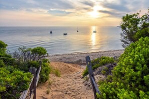 A beach with a wooden fence leading to the shore, a sandy path, and boats on the water. - Gorgeous Architectural Waterfront Property! (Eastham)
