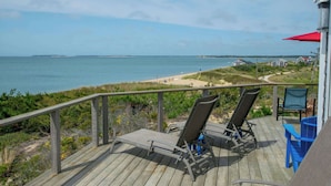 Eine Holzterrasse mit zwei Liegestühlen und einem blauen Tisch, mit Blick auf einen Strand und den Ozean.