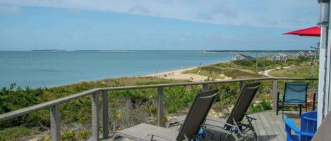 A wooden deck with two lounge chairs and a blue table, overlooking a beach and ocean.