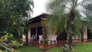 A covered outdoor seating area with chairs and tables, surrounded by lush greenery and a palm tree. - Comfortable house (large balcony and garden), 70 meters from the beach with Wi-Fi.   (praia da lagoinha ubatuba)