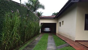 A pathway leading to a car parked under a covered area, surrounded by a hedge and a building with a tiled roof. - Comfortable house (large balcony and garden), 70 meters from the beach with Wi-Fi.   (praia da lagoinha ubatuba)