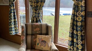 Une pièce chaleureuse avec un fauteuil à motifs floraux, un coussin orné d’un motif de chat et une vue sur un lac, visible à travers des rideaux à motifs.