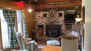 Un salon chaleureux avec une cheminée en briques, des poutres apparentes, un téléviseur et un coin repas comprenant une table et des chaises.