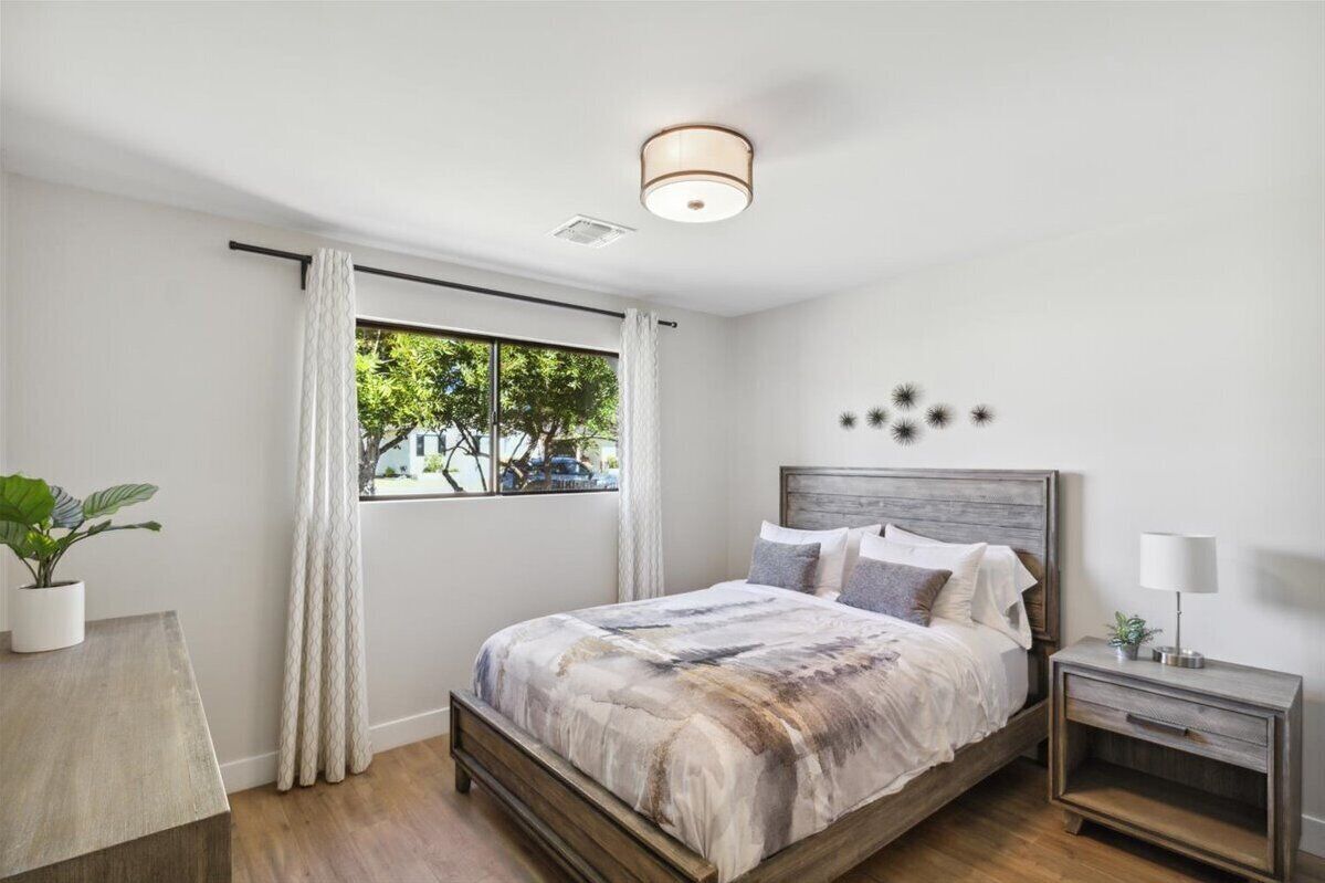 A bedroom with a wooden bed, a bedside table with a lamp, a window with curtains, and a view of trees outside.