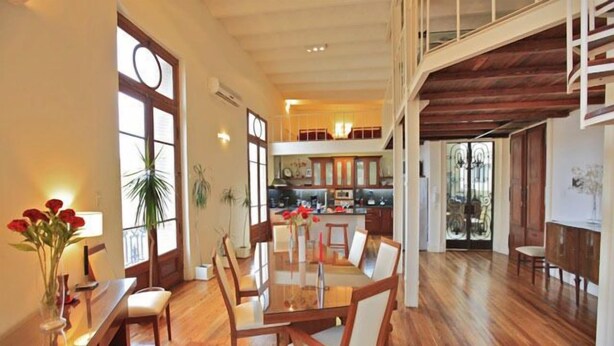 Un vaste espace à manger avec une table et des chaises en bois, de grandes fenêtres et une vue sur la cuisine et l’étage supérieur.