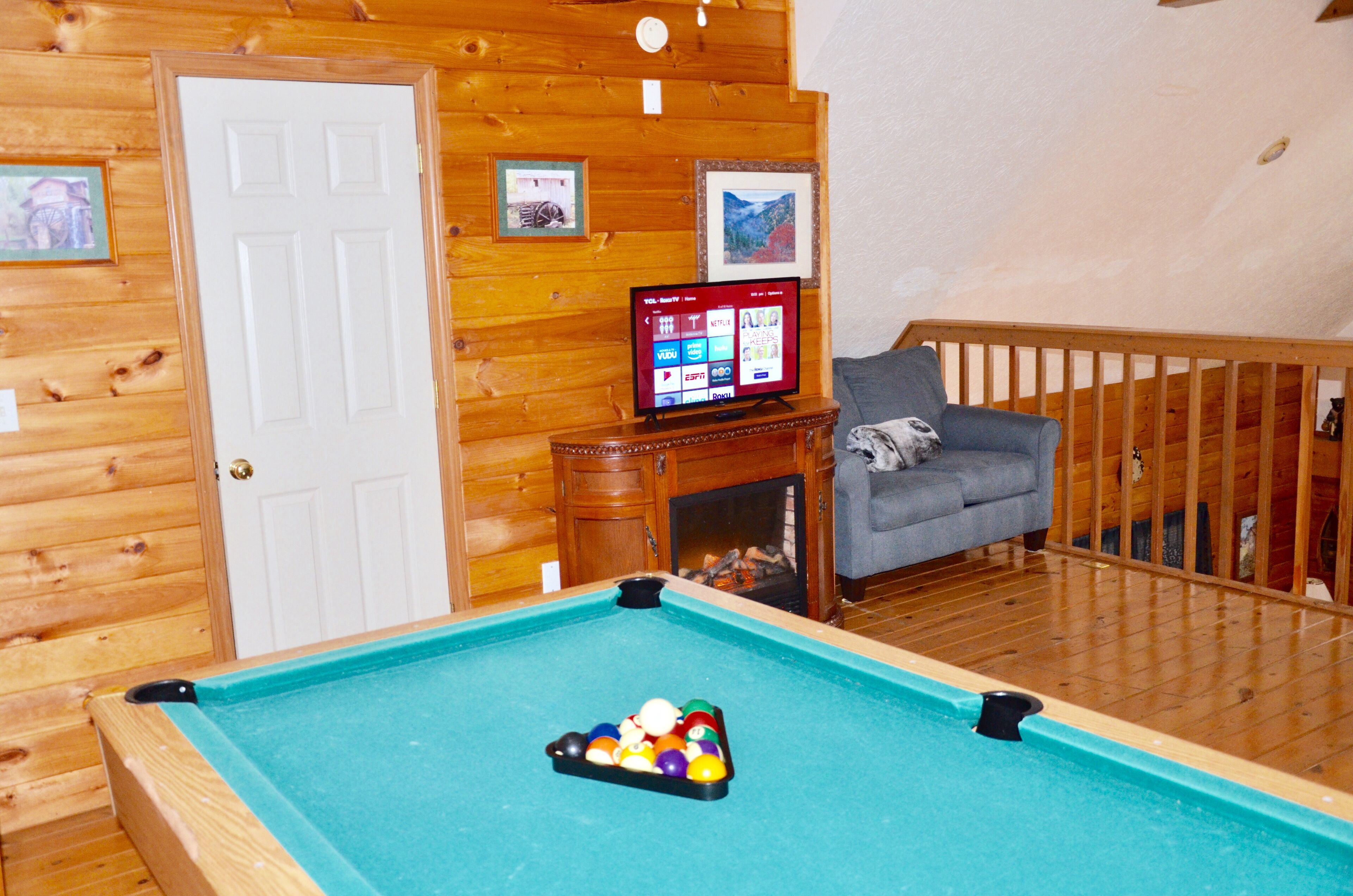 A pool table with balls, a television displaying a streaming service interface, a grey sofa, and wooden walls.