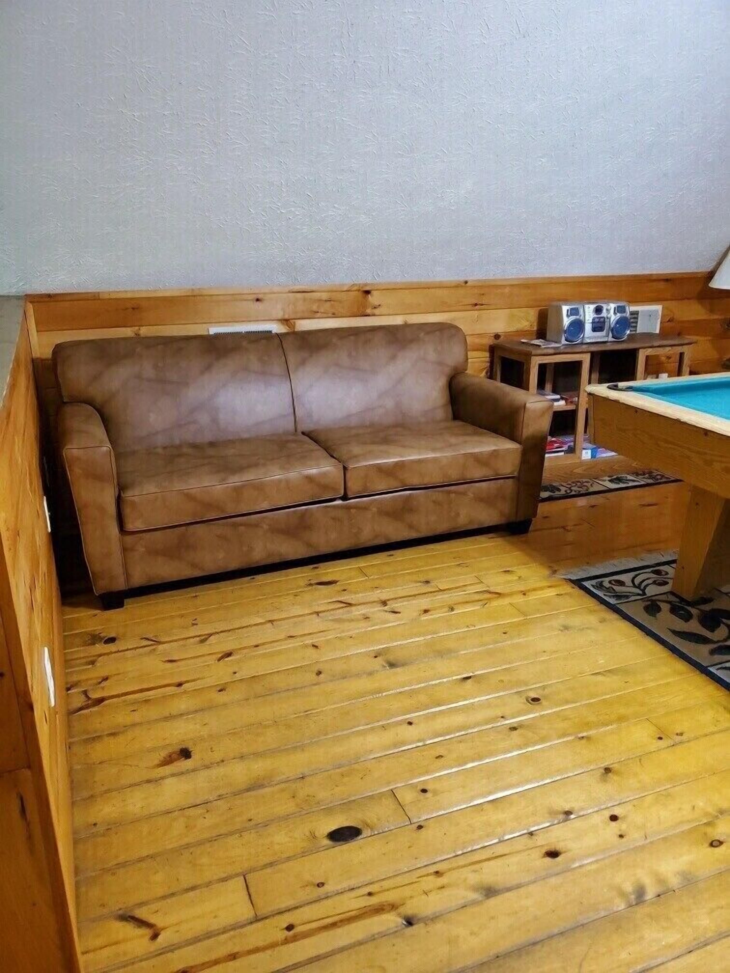 A wooden-floored room with a brown leather sofa, a pool table, and a small wooden cabinet with speakers.