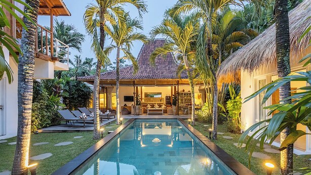 Ein Pool mit freiem Blick auf die Umgebung, darunter ein Haus mit Ziegeldach und einer Struktur mit Strohdach, umgeben von Palmen und einem gepflegten Rasen.