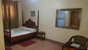 A room with a wooden four-poster bed, a small bedside table with a phone, a chair with a menu, and a mirror on the wall. - Hotel Hensek (Kampala)
