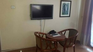 A room with a flat-screen TV mounted on the wall, two wooden chairs with cushioned seats, a small table, and a framed picture on the wall. - Hotel Hensek (Kampala)