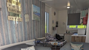 Un espacio de sala acogedor con un sofá, una mesita y un diván. Hay una zona de cocina con un microondas y un fregadero. La habitación tiene suelo de madera y una pared con un letrero decorativo.