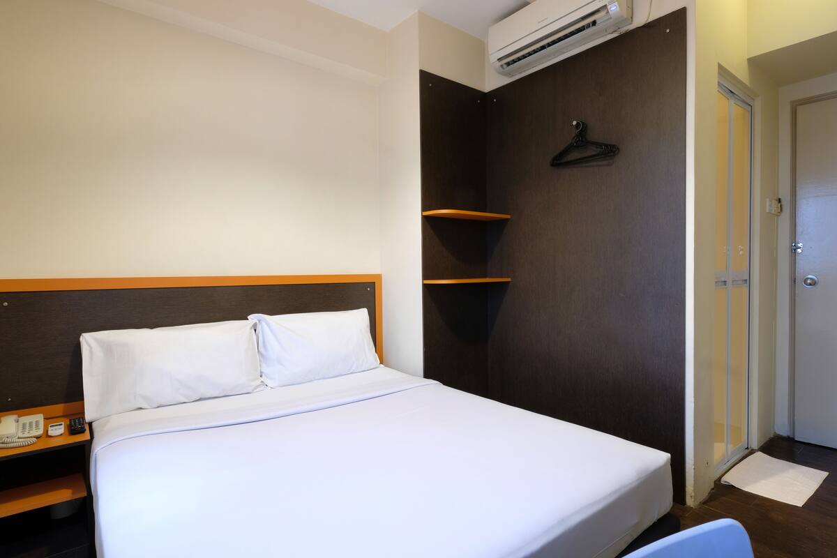 A hotel room with a bed, a bedside table with a phone, a wall-mounted air conditioner, and a door with a mat.