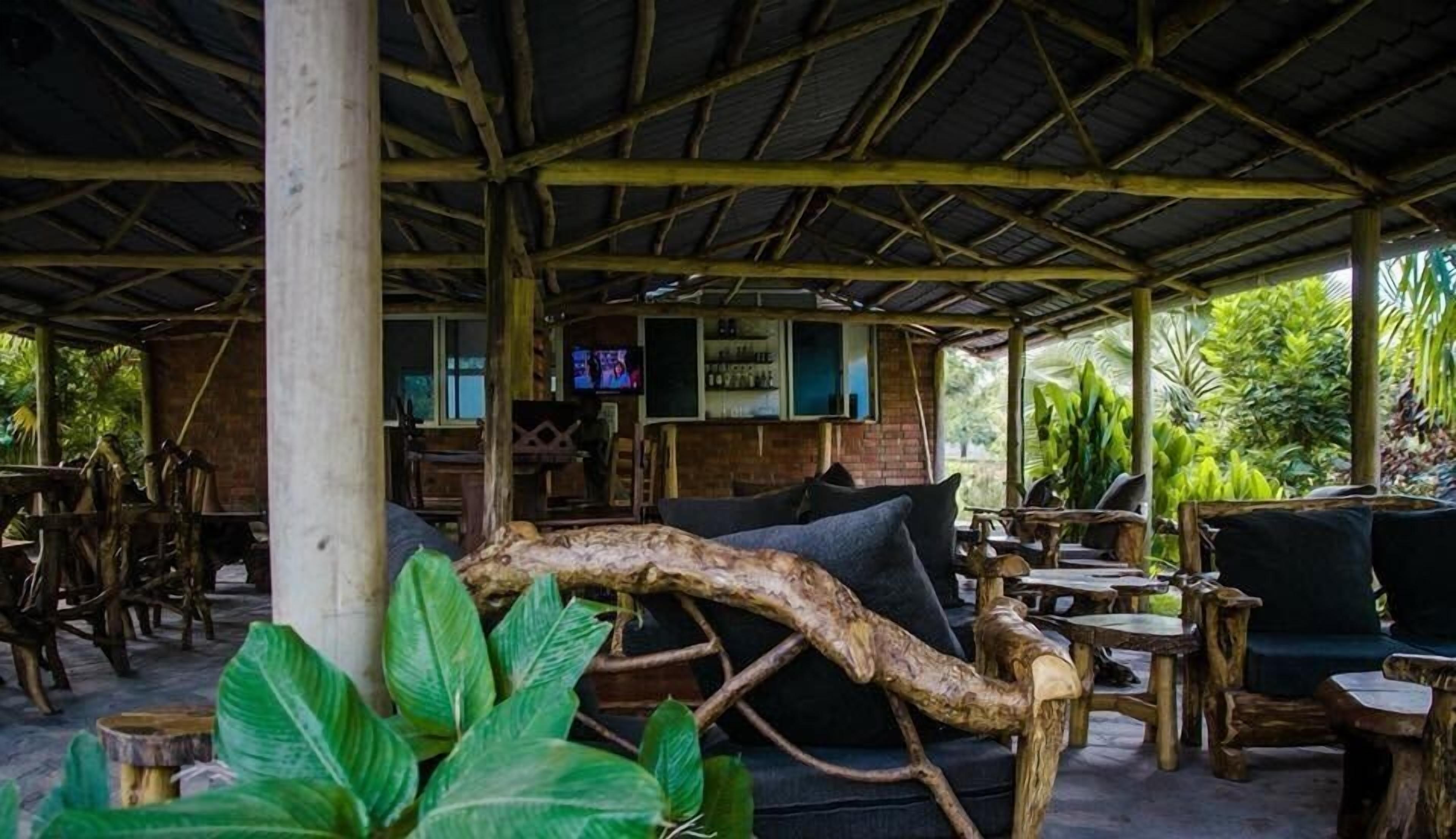 An outdoor seating area with rustic wooden furniture, a large tree branch used as a decorative element, and a brick wall with a television screen.